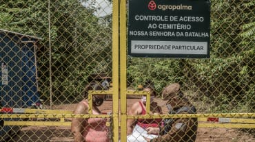 Duas mulheres sendo controladas por um guarda por trás de um portão gradeado - Texto inscrito na placa da empresa: Agropalma - Controle de Acesso ao Cemitério Nossa Senhora da Batalha. PROPRIEDADE PARTICULAR