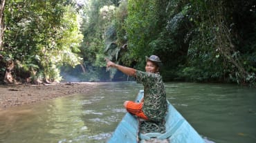 Um homem na ponta de um pequeno barco mostra, entusiasmado, a floresta tropical