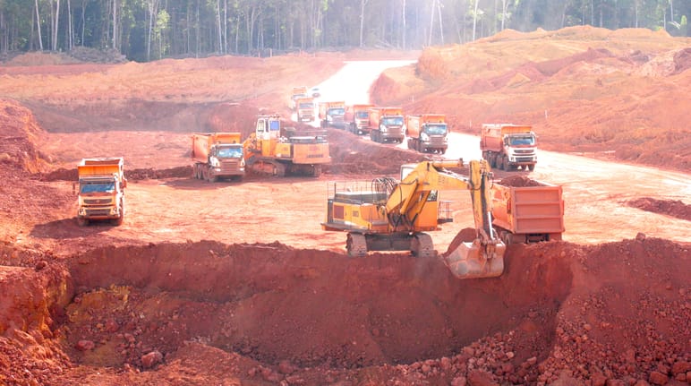Uma escavadeira empurra o solo vermelho de um poço para dentro de um caminhão. Atrás dela, uma fila de caminhões de espera e máquinas de construção é visível. Árvores na floresta tropical em segundo plano