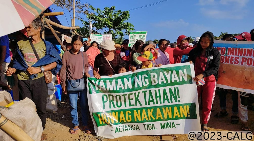 Grupo de pessoas segura um cartaz, no qual se lê (traduzido): “Protejam a riqueza do povo! Nada de roubo! Aliança contra a mineração
